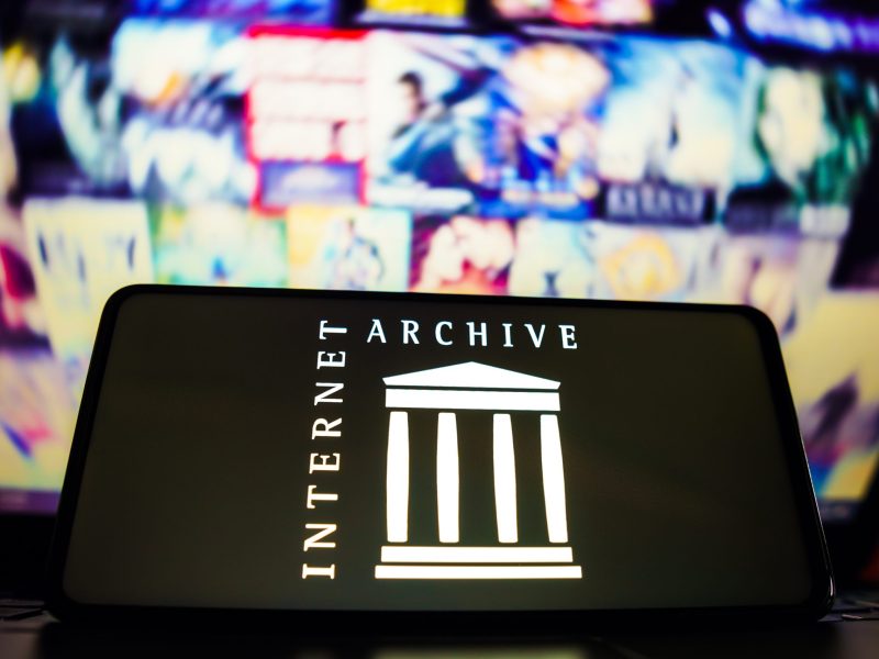 The Internet Archive logo, which resembles the entrance to a columned building, is displayed a phone screen. In the background are a series of screens displayed in a grid with technicolor, out of focus images playing.