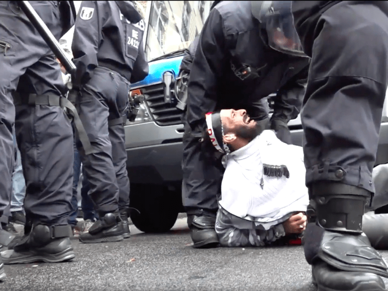 A Middle Eastern man in all white lays down on the pavement with his head rolled back. He is crying out in pain as a black-uniformed police officer bends over to restrain him. In the frame are the black pants and boots of three other cops.